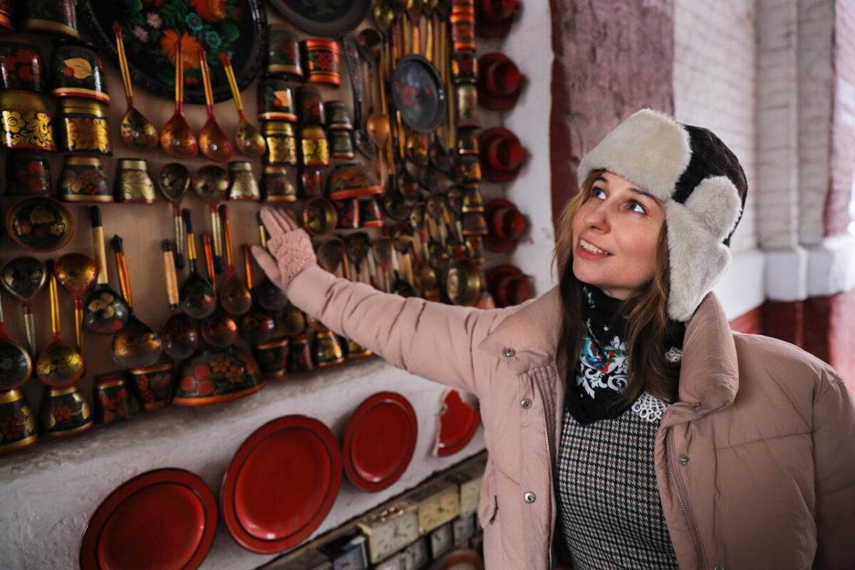 Woman in winter clothing admiring traditional Russian decorated wall indoors.